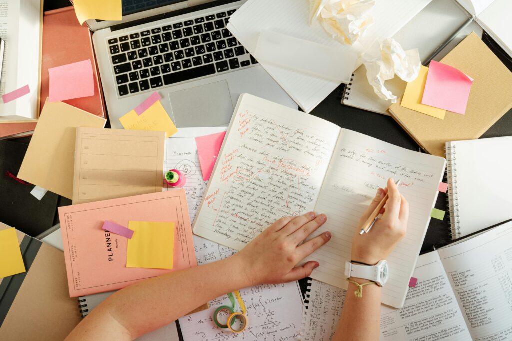 Top-down view of a student's hands writing in a notebook on a messy desk covered with sticky notes, textbooks, and a laptop, illustrating the need for assignment help for students.