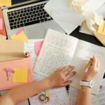 Top-down view of a student's hands writing in a notebook on a messy desk covered with sticky notes, textbooks, and a laptop, illustrating the need for assignment help for students.