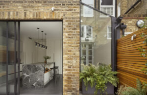 Exterior view of a modern glass and brick house extension in London, featuring large sliding doors opening into a contemporary kitchen with a dark marble island.
