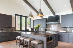 A modern, high-end kitchen within a spacious house addition featuring vaulted ceilings with exposed beams, charcoal cabinetry, and a large central island, demonstrating the luxury potential of home extensions.