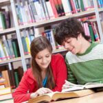 Teenage girl and boy happily study from an open textbook together in a library for GCSE resit revision, with books on the table and shelves in the background.