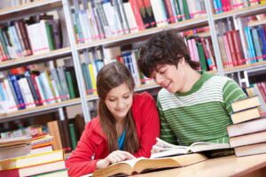 Teenage girl and boy happily study from an open textbook together in a library for GCSE resit revision, with books on the table and shelves in the background.