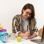 smiling-young-student-girl-wearing-glasses-sitting-desk-with-university-tools-using-mobile-phone-isolated-white-wall (1) (1) (1) (1) (1) (1)